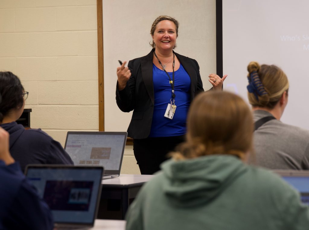Jessica Roisen is shown lecturing in front of a group of students. She is wearing a blue shirt and blazer and is smiling.