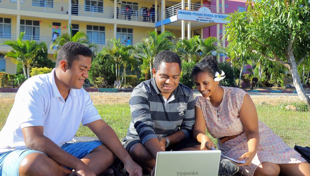 Three students sit outside of a building on the University of Fiji's campus.